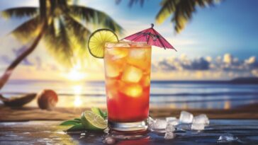 Tropical tequila cocktail with ice, lime slice, and a pink umbrella, set on a beachside table with a sunset, palm trees, and ocean in the background.